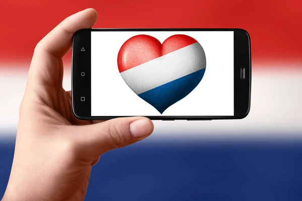 전화 화면에 심장 모양으로 네덜란드 국기가 걸려 있습니다. 손에 들고 있는 스마트폰에 하트 플래그가 보 입니다.. — 스톡 사진
