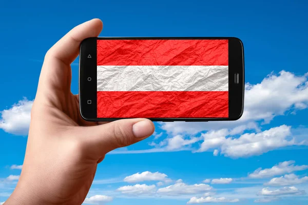 Austria flag on the phone screen. Smartphone in hand shows a flag on a background of the sky with clouds.