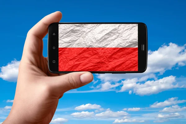 Bandera de Polonia en la pantalla del teléfono. Smartphone en la mano muestra una bandera en un fondo del cielo con nubes . —  Fotos de Stock