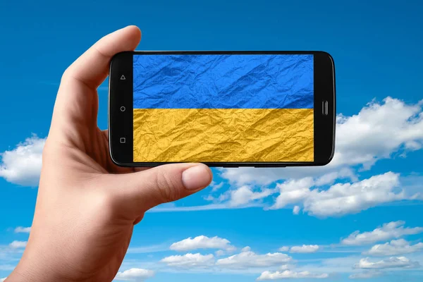 Флаг Украины на экране телефона. Смартфон в руке показывает флаг на фоне неба с облаками . — стоковое фото