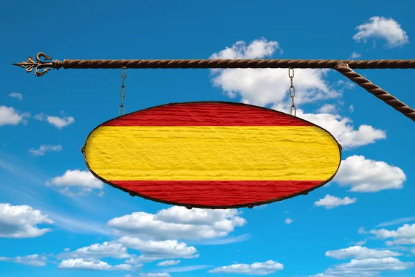 Spain flag on a signboard. Oval sign in the colors of the flag Spain hanging on a metal forged design. Template on a background of blue sky with clouds.