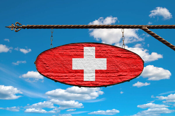 Switzerland flag on signboard. Oval signboard colors flag Switzerland hangs on a metal forged structure. Template on a background of blue sky with clouds. 
