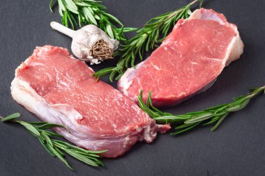 Çiğ et. İki dilim taze çiğ et, bir dal biberiye ve sarımsak siyah bir taş tahtada. Barbekü için et hazırlıyorum. Sağlıklı, doğal yiyecekler..