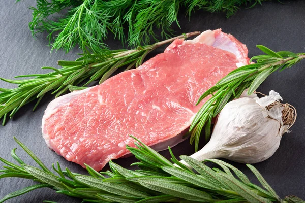Raw meat. Slice of fresh raw meat, rosemary and garlic on a black slate stone board. Preparing meat for a barbecue. Healthy, natural food.