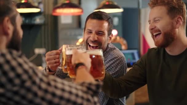 パブでビールマグカップをクリックして幸せな中大人の友人 3人の陽気な男がドラフトビールを飲み 会議や笑顔を祝う バーで夜の間に冷たいビールを楽しんでいる若い男性を笑って — ストック動画