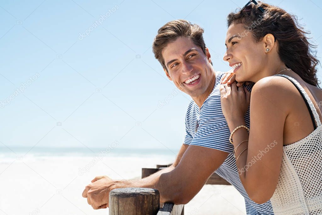 Loving couple standing at beach shore while looking away with copy space. Handsome young man looking at beautiful woman with love on beach while she is contemplating the future. Boyfriend and girlfriend enjoying summer vacation at the ocean with copy