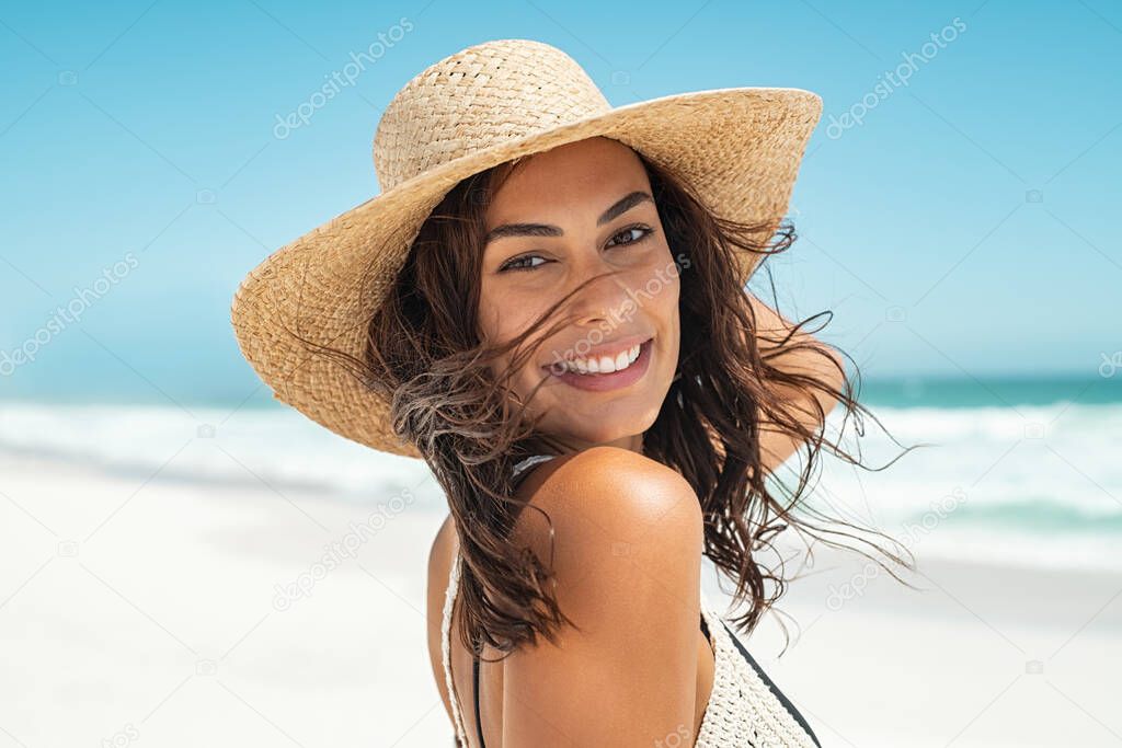 Portrait of beautiful smiling young woman wearing straw hat at beach with sea in background. Beauty fashion girl looking at camera at seaside with big grin. Carefree tanned woman walking on sand and laughing.