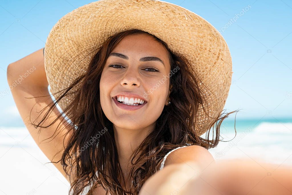 Portrait of beautiful young woman in casual wearing straw hat at seaside. Cheerful young woman smiling at beach during summer vacation. Happy girl with black hair and freckles enjoying the sun.