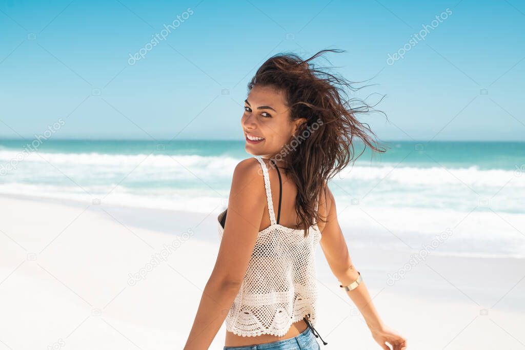 Beautiful latin woman on tropical beach running away while looking at camera. Portrait of happy tanned woman smiling with sea in background. Fashionable girl in casual clothing enjoying and running on tropical beach with copy space.