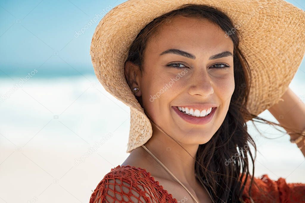 Portrait of young beauty woman with ocean in background. Beautiful fashion woman at the beach protecting herself from the sun with a big hat and copy space. Close up face of glamour girl with sunhat at sea looking at camera.