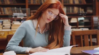 Gözlüklü kadın kütüphanede oturup kitap okuyor. Eski kütüphanede konsantrasyonla çalışan üniversite öğrencisinin portresi. Kızıl saçlı kız üniversite kütüphanesinde oturuyor ve ödevlerini yapıyor..