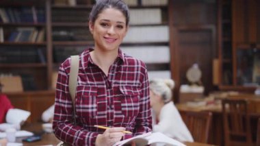 Sınıf arkadaşları arka planda çalışırken mutlu bir üniversite öğrencisi kameraya bakıyor. Elinde not ve kalemle kolej kütüphanesinde duran gülümseyen genç bir kadının portresi. Üniversitede okuyan kendinden emin Latin kız..