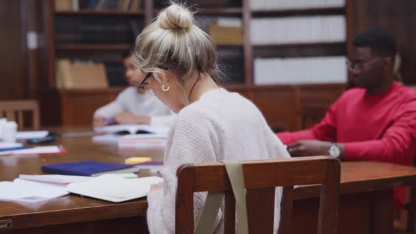 在图书馆学习的女大学生的肖像 快乐的年轻女子戴着眼镜在大学图书馆学习 看着相机 满意的女孩在为考试学习 — 图库视频影像