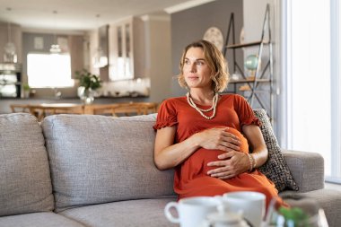 Düşünceli orta yaşlı hamile kadın kanepeye oturmuş fotokopi odasıyla başka yere bakıyor. Annemin evdeki kanepede dinlenmesini bekliyorum. Bebek karnında elleri olan güzel hamile kadın doğmamış çocuğu düşünüyor..