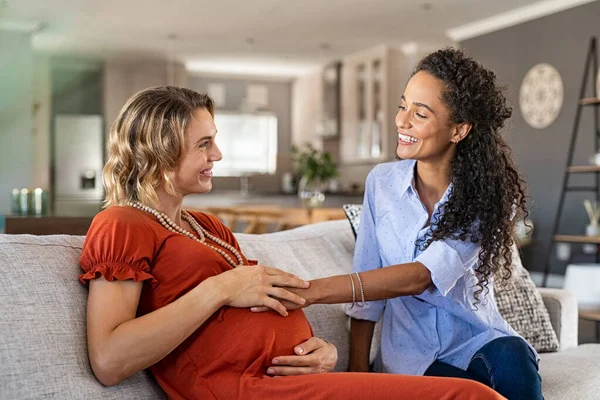 快乐的非洲裔美国朋友感觉到孕妇的肚子痛 快乐的女孩手牵着手期待着妈妈的胃摸着宝宝的动作 年轻女子坐在沙发上和朋友说话 摸着肚子 — 图库照片