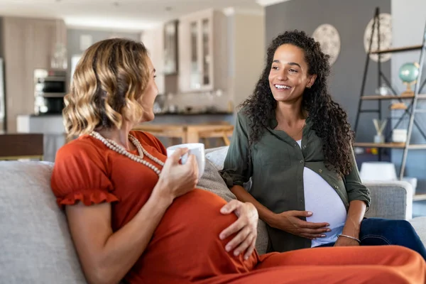 两名多族裔孕妇在家里喝茶 笑着孕妇说话 互相看着对方 快乐的女孩们 带着宝宝在客厅里放松一下 并进行友好的交谈 — 图库照片