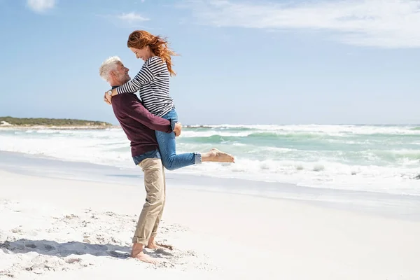 在阳光普照的日子里 一对成熟的夫妇在沙滩上拥抱 相得益彰 快乐的妻子在海滩上拥抱她的丈夫 双脚离开地面 亲爱爱老人爱老人爱老人 老年人 老年人 老年人在老年人 — 图库照片