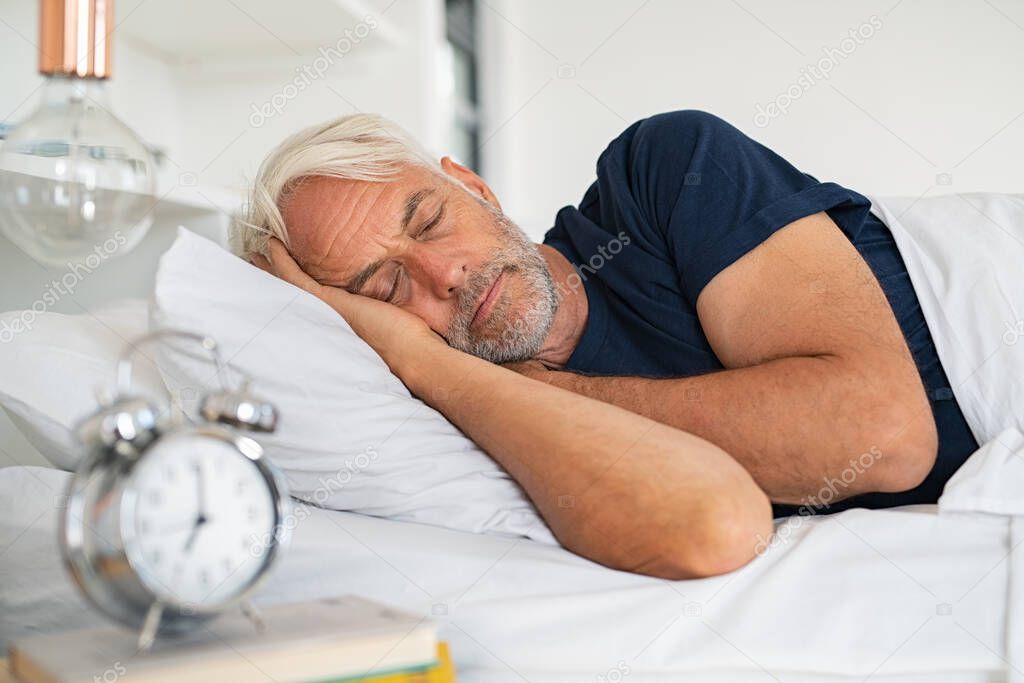 Old tired man sleeping on bed at home. Senior man with white hair in deep sleep on soft pillow at home. Mature healthy guy lying on side resting at home while sleeping.