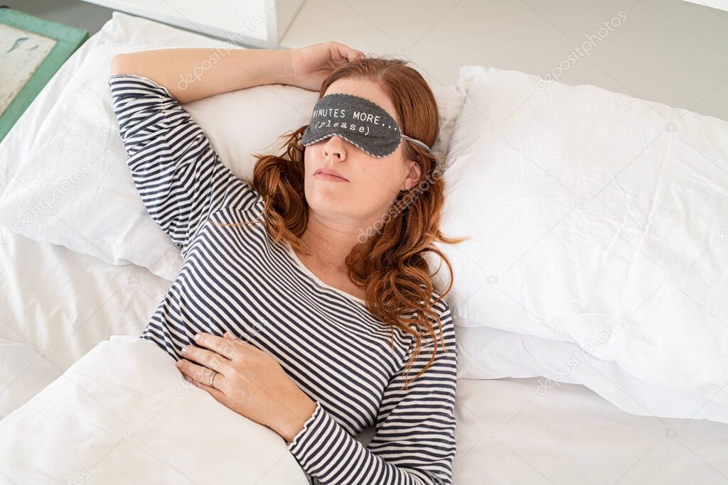 Top view of mature woman with an eye mask sleeping on bed. Beautiful sleepy middle aged woman wearing funny eye mask. High angle view of redhead woman in pajamas dreaming on white bed with eye mask with five minutes more text.