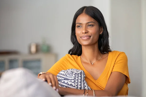 Young indian woman relaxing on couch at home while looking away. Thoughtful smiling mixed race girl daydreaming while sitting on sofa at home. Pensive middle eastern woman thinking.