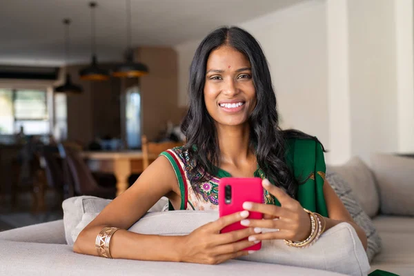 伝統的なサリの笑顔インド人女性の自宅でスマートフォンを使用しての肖像画 携帯電話を持ちながらソファに座ってカメラを見ている若いインド人女性 美しいですHindu妻とともにBindiサーフィンネットとともに携帯電話と見ますA — ストック写真