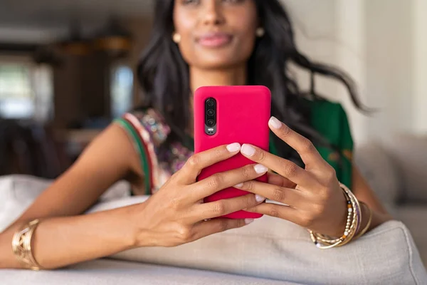 Close up hands of young indian woman in traditional clothing holding smartphone. Hindu girl in ethnic wear, sari, with bracelets sitting on couch and using cellphone. Detail of indian girl hands typing message on mobile phone at home.