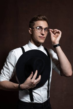 Yuvarlak gözlüklü, askılı, beyaz gömlekli ve şapkalı bir adam..