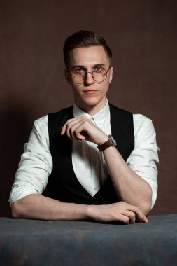 Beyaz gömlekli, siyah yelekli ve yuvarlak gözlüklü bir adam masada oturuyor.