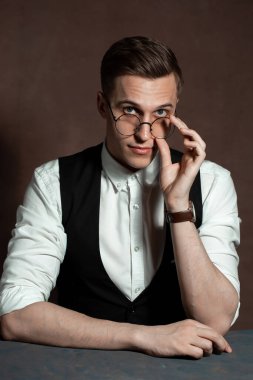 Beyaz gömlekli, siyah yelekli ve yuvarlak gözlüklü bir adam masada oturuyor.