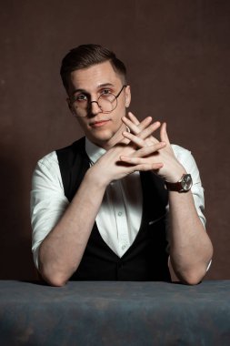 Beyaz gömlekli, siyah yelekli ve yuvarlak gözlüklü bir adam masada oturuyor.