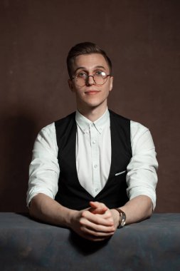 Beyaz gömlekli, siyah yelekli ve yuvarlak gözlüklü bir adam masada oturuyor.