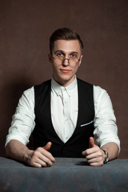 Beyaz gömlekli, siyah yelekli ve yuvarlak gözlüklü bir adam masada oturuyor.