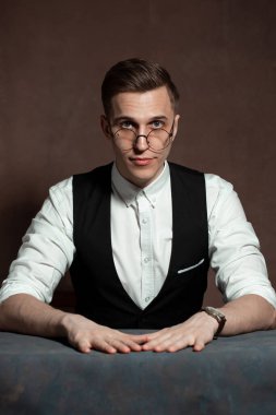 Beyaz gömlekli, siyah yelekli ve yuvarlak gözlüklü bir adam masada oturuyor.