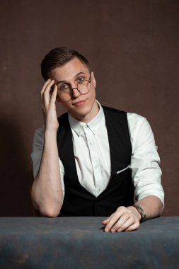 Beyaz gömlekli, siyah yelekli ve yuvarlak gözlüklü bir adam masada oturuyor.