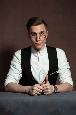 Beyaz gömlekli, siyah yelekli ve yuvarlak gözlüklü bir adam masada oturuyor.