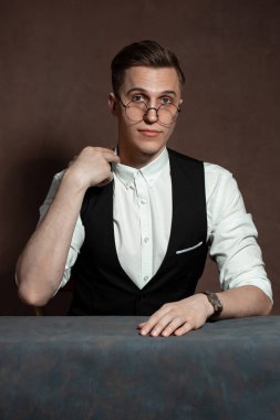 Beyaz gömlekli, siyah yelekli ve yuvarlak gözlüklü bir adam masada oturuyor.