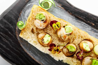 Sarımsak ve peynir kaplı salyangozlar. Soslu leziz salyangozlar. Ahşap tepside yemek tabağı. Ekmeğin üstünde menekşelerle servis edilen lezzetli bir yemek. Fransız yemek sunumu, mutfak menüsü