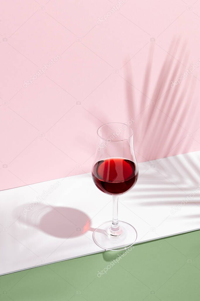 Red wine on creative background. Shadows of palm leaf on pink walls and white floor. Pink, white and green. 