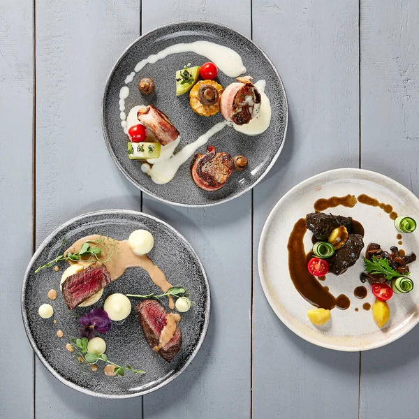 Hot meals, meat appetizers in ceramic plates top view. Veal cheeks, pork in bacon, Veal tenderloin filet mignon. Meat with vegetables and sauces. Gastronomy, served restaurant dishes on wooden table