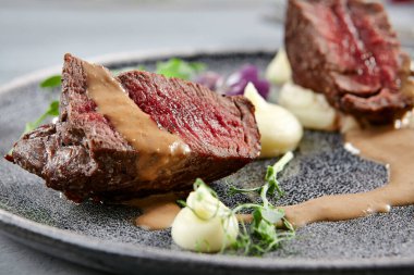Kök ekili kremalı dana fileto mignon gri tabakta. Soslu sığır eti dilimleri, yeşillik ve menekşe çiçekleri yakın plan. Et aperatifleri, restoran yemekleri, lezzetli yemekler.