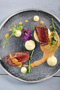 Kök ekili kremalı dana fileto mignon gri tabakta. Soslu sığır eti dilimleri, yeşillik ve menekşe çiçekleri yakın plan. Et aperatifleri, restoran yemekleri, lezzetli yemekler.