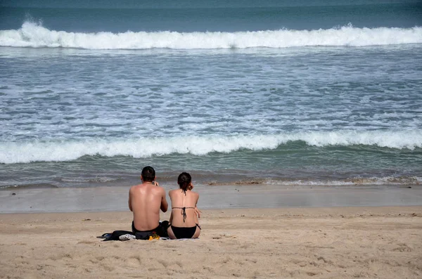 Mladý pár sedí na bílém písku, vidět na moři surfovat s pěnou v Bal — Stock fotografie