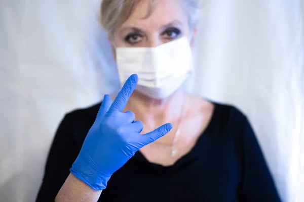 Senior woman wearing mask and wearing gloves shows the rock sign. Coronavirus and epidemic virus symptoms.