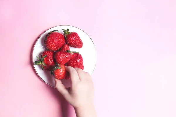 上からの眺めピンクの背景にイチゴを手に取る子供たち イチゴのプレート 夏の健康的な食事のコンセプト — ストック写真