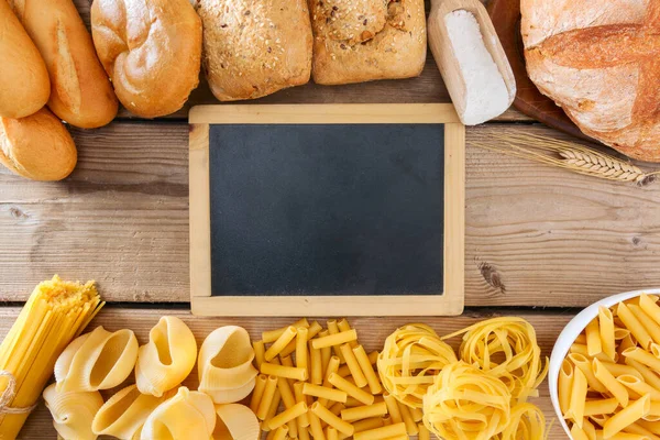 Various types of bread and pasta with  in the middle is a blackboard with copy space photographed on a wooden table in flat lay.