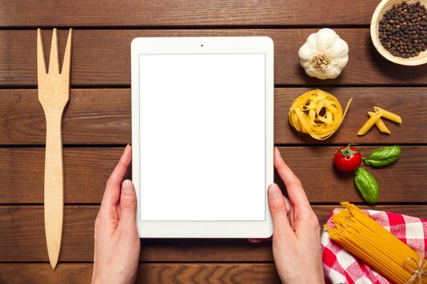 Female hands holding a white tablet with copy space on a wooden background with ingredients for cooking Italian pasta, top view