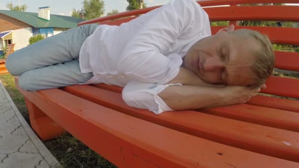 Мужчина в джинсах и рубашке спит на скамейке под открытым небом — стоковое видео