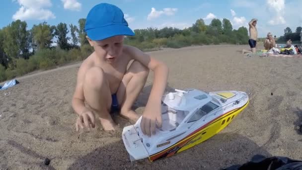 Ufa, russland - 28. Juli 2016: Ein siebenjähriger Junge mit blonden Haaren startet ein ferngesteuertes Boot auf dem Fluss. Strand, Sommer, Fluss, Russland — Stockvideo