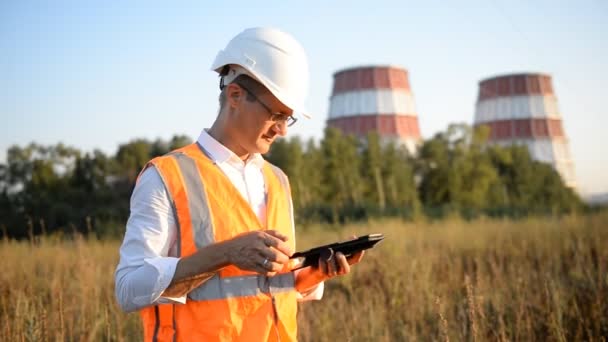 A man (engineer, worker, specialist) in a helmet and waistcoat, with a tablet on the background of an industrial landscape — Stock Video