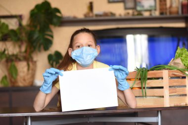 Genç bir kadın boş levhayı tutuyor. Coronavirus. Yüz maskesi takmış bir kadın, Coronavirus krizi sırasında mutfakta aşçılık yapıyor. Evde kal. Evde yemek pişirmenin tadını çıkar. Aile kavramı.